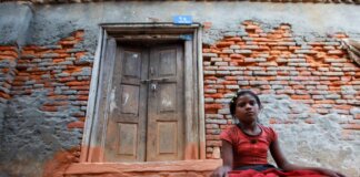 A young girl sits calmly by a rustic brick wall with a wooden doorway, conveying resilience and tranquility.