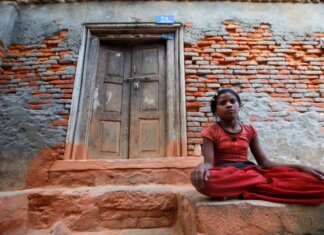 A young girl sits calmly by a rustic brick wall with a wooden doorway, conveying resilience and tranquility.