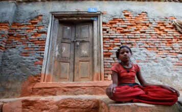 Детская бедность в Канаде удвоилась с 2020 года — доклад ЮНИСЕФ A young girl sits calmly by a rustic brick wall with a wooden doorway, conveying resilience and tranquility.