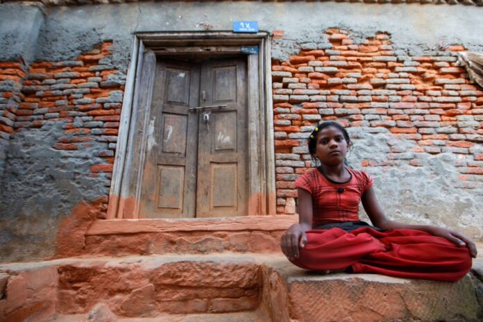 A young girl sits calmly by a rustic brick wall with a wooden doorway, conveying resilience and tranquility. A young girl sits calmly by a rustic brick wall with a wooden doorway, conveying resilience and tranquility.