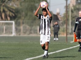 A young soccer player prepares to throw-in during a match on a sunny day, with referee nearby.