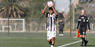 A young soccer player prepares to throw-in during a match on a sunny day, with referee nearby.