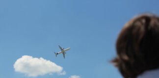 An airplane flies high in a clear sky while a person watches from below.