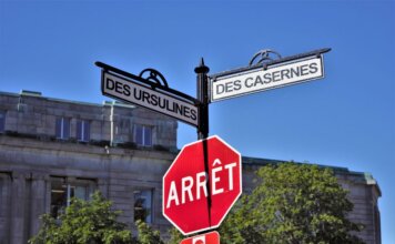 Bilingual street signs with stop sign against a clear blue sky in a city setting, featuring historical street names.