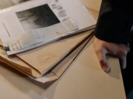 Close-up of a hand resting on a desk near a stack of investigative documents and newspaper clippings.