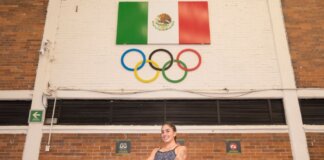 Confident female athlete in swimsuit poses below Mexican flag and Olympic rings indoors.