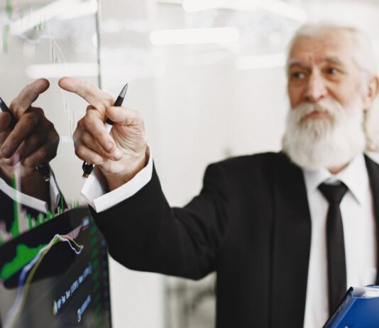 Elderly businessman with beard presenting financial trends on a screen.