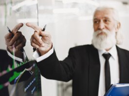 Elderly businessman with beard presenting financial trends on a screen.
