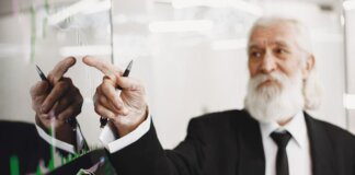 Elderly businessman with beard presenting financial trends on a screen.
