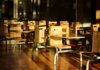 Empty wooden chairs and tables in a sunlit cafeteria setting, casting dramatic shadows.