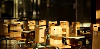 Empty wooden chairs and tables in a sunlit cafeteria setting, casting dramatic shadows.