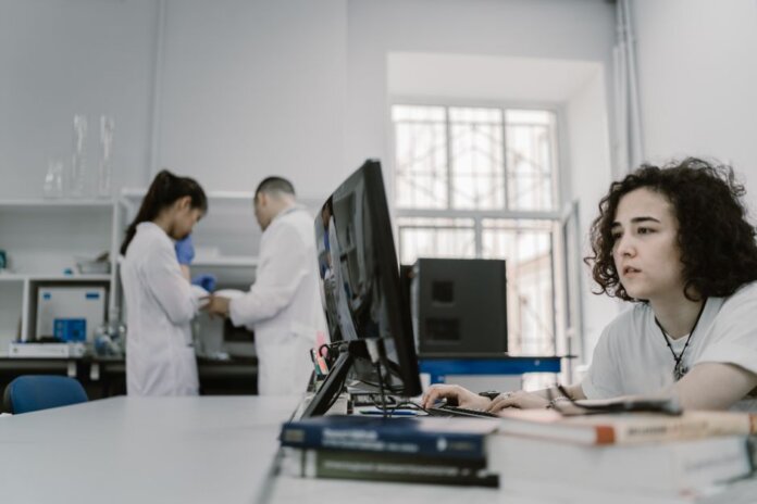 Focused woman working on a computer in a busy laboratory setting, showcasing teamwork and scientific research. Focused woman working on a computer in a busy laboratory setting, showcasing teamwork and scientific research.
