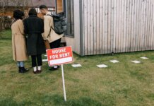 Group of adults viewing a wooden house with a 'House for Rent' sign on the lawn.