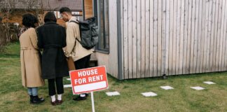 Group of adults viewing a wooden house with a 'House for Rent' sign on the lawn.