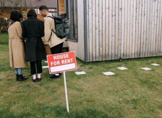 Group of adults viewing a wooden house with a 'House for Rent' sign on the lawn.