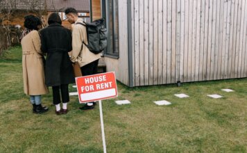 Арендодатель обвиняется в установке скрытых камер в съёмных домах Group of adults viewing a wooden house with a 'House for Rent' sign on the lawn.