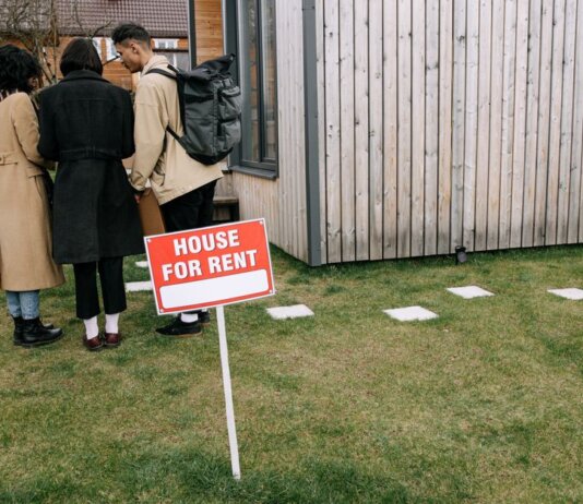 Group of adults viewing a wooden house with a 'House for Rent' sign on the lawn.