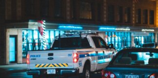 Police pickup with tail light riding on asphalt roadway near car along building with glass showcase in city on evening time