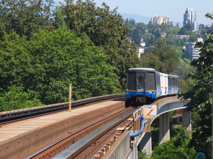 SkyTrain travels on elevated tracks through New Westminster, surrounded by lush greenery.