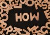 Top view of scattered wooden letters with 'HOW' centered on a dark surface, promoting inquiry.