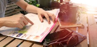 Traveler pointing at map on wooden table with camera and accessories under sunlight.