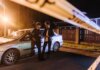 Two men discussing at a nighttime urban crime scene with police tape and a car.