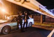 Two men discussing at a nighttime urban crime scene with police tape and a car.