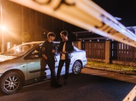 Two men discussing at a nighttime urban crime scene with police tape and a car.