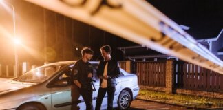 Two men discussing at a nighttime urban crime scene with police tape and a car.