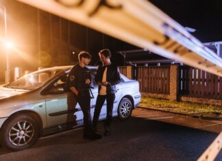 Two men discussing at a nighttime urban crime scene with police tape and a car.