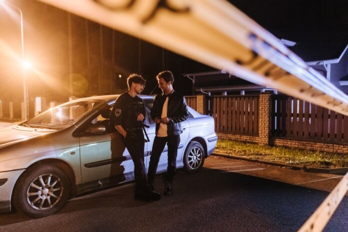 Two men discussing at a nighttime urban crime scene with police tape and a car.