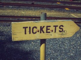 Weathered wooden ticket sign pointing towards railway tracks outdoors, vintage look.