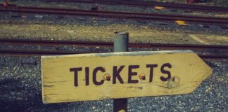 Weathered wooden ticket sign pointing towards railway tracks outdoors, vintage look.