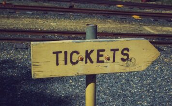 Weathered wooden ticket sign pointing towards railway tracks outdoors, vintage look.