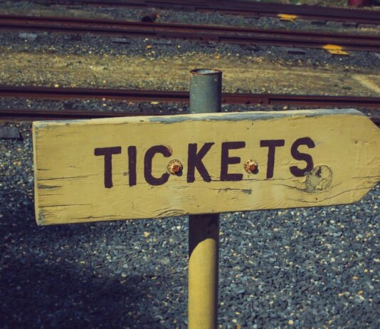 Weathered wooden ticket sign pointing towards railway tracks outdoors, vintage look.
