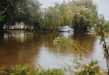 Канада выделит экстренную помощь Манитобе для борьбы с наводнением A car struggles through a flooded street surrounded by trees after a heavy rainstorm.