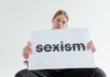 A caucasian woman confidently holds a sign addressing sexism in a bright studio setting.