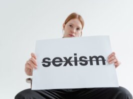 A caucasian woman confidently holds a sign addressing sexism in a bright studio setting.