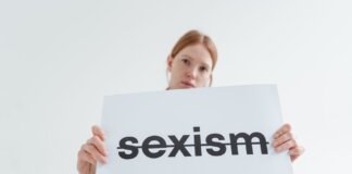 A caucasian woman confidently holds a sign addressing sexism in a bright studio setting.