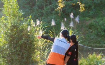 A humanitarian worker wearing a World Vision vest assists a child in a lush, sunny environment.