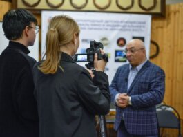 A journalist records an interview with a man using a camcorder indoors.