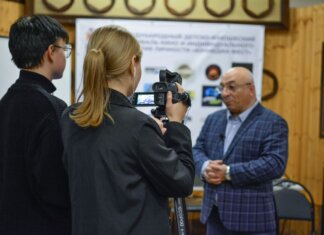A journalist records an interview with a man using a camcorder indoors.