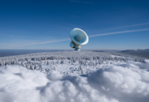 A large satellite dish observatory surrounded by snowy landscape and winter forest under a clear blue sky.