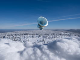 A large satellite dish observatory surrounded by snowy landscape and winter forest under a clear blue sky.