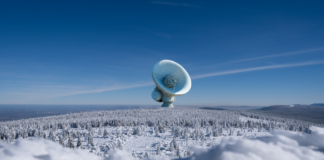 A large satellite dish observatory surrounded by snowy landscape and winter forest under a clear blue sky.