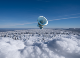 A large satellite dish observatory surrounded by snowy landscape and winter forest under a clear blue sky.