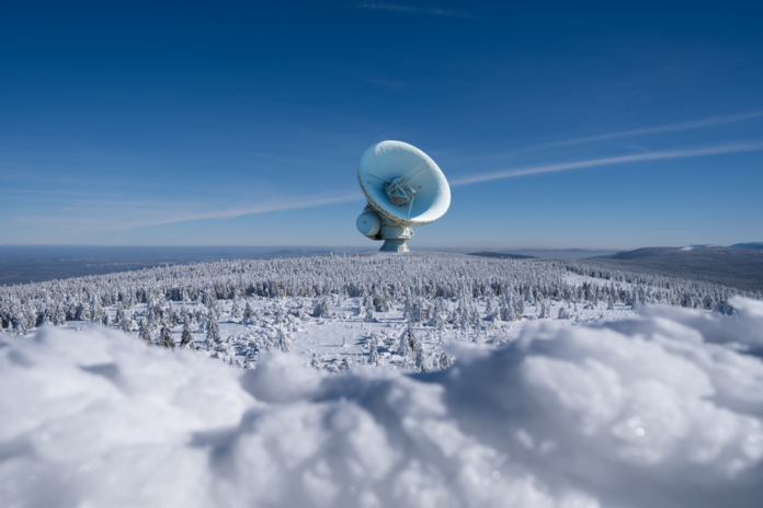 A large satellite dish observatory surrounded by snowy landscape and winter forest under a clear blue sky.
