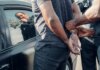 A man being handcuffed by police near a car, with reflection visible.