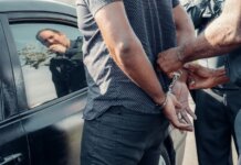 A man being handcuffed by police near a car, with reflection visible.