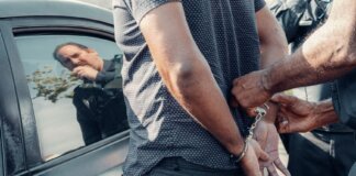 A man being handcuffed by police near a car, with reflection visible.
